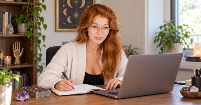 A red-haired psychic reader writes profile notes beside a laptop, tarot deck and amethyst.